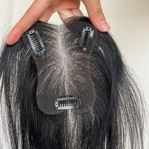 Könnte beinhalten: Ein schwarzes Haarteil mit einer feinen Netzbasis und drei schwarzen, druckempfindlichen Metallclips an der Unterseite. Das Haar ist glatt und dunkel und erstreckt sich von dem zentralen Netzeinsatz, der das Aussehen einer natürlichen Kopfhaut imitiert.