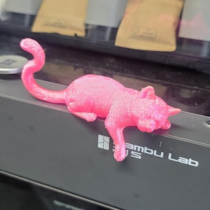 Może przedstawiać: Jasnoróżowa figurka kota wydrukowana w 3D, leżąca ze zwiniętym ogonem. Kot spoczywa na czarnej powierzchni z napisem "Bambu Lab". W tle widać drukarkę 3D.