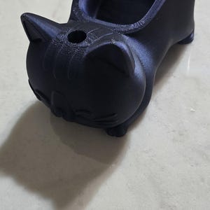 May include: A dark blue cat-shaped planter. The planter has a rounded body, a small tail, and a hole in the top. The cat's face has small ears and a smiling expression. The planter is made of a 3D-printed material.