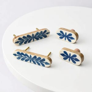 May include: Two long rectangular wooden drawer pulls and two small oval wooden knobs, each featuring a white inlay with a dark blue painted botanical leaf pattern and gold-toned metal hardware for mounting to furniture surfaces.