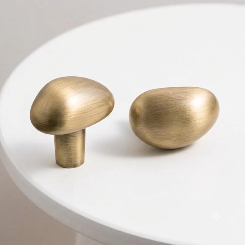 Op de afbeelding: Twee kastknoppen met een geborstelde messing afwerking en een organische, kiezelsteenachtige vorm. E&eacute;n knop staat rechtop met de cilindrische basis zichtbaar, terwijl de andere op zijn kant ligt, wat de gladde, afgeronde textuur benadrukt.