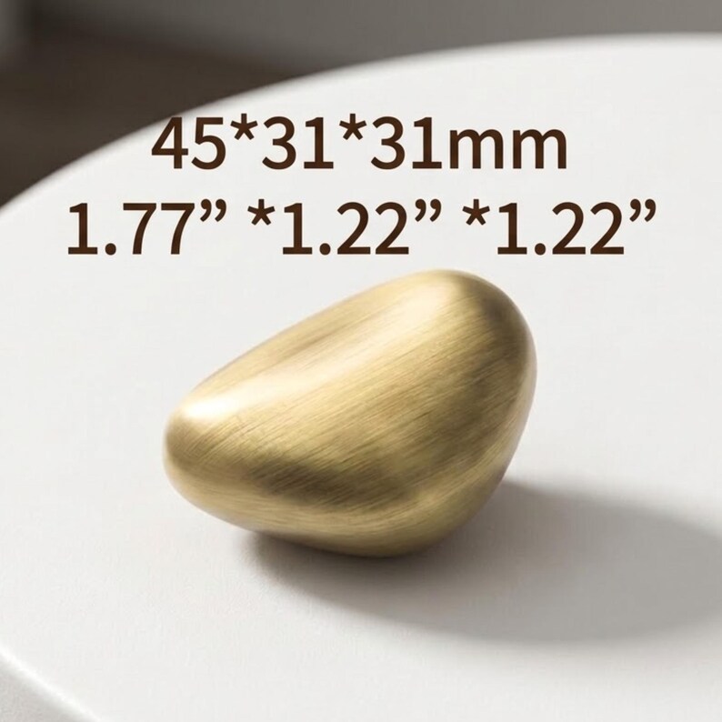 Op de afbeelding: Een geborstelde goudkleurige meubelknop met een onregelmatige, kiezelachtige vorm. De afmetingen staan vermeld als 45*31*31mm en 1.77" * 1.22" * 1.22". Het oppervlak heeft een gladde, metalen afwerking met een zichtbare horizontale korrel.