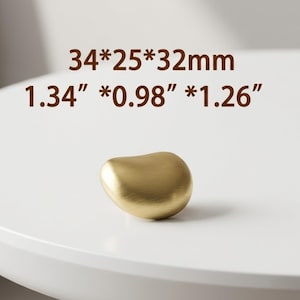 Op de afbeelding: Een goudkleurige meubelknop met een gladde, afgeronde, kiezelsteenachtige vorm en een geborstelde metalen afwerking. De tekst 34*25*32mm en 1.34*0.98*1.26 inches staat boven het object gedrukt en geeft de fysieke afmetingen aan.
