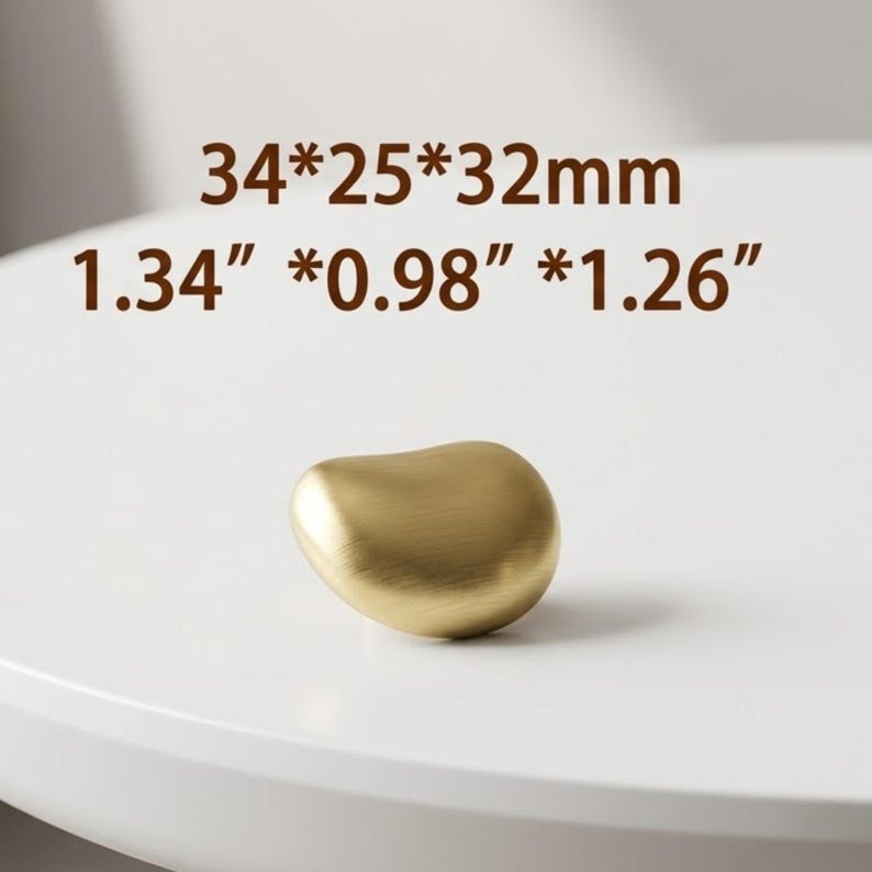 Op de afbeelding: Een goudkleurige meubelknop met een gladde, afgeronde, kiezelsteenachtige vorm en een geborstelde metalen afwerking. De tekst 34*25*32mm en 1.34*0.98*1.26 inches staat boven het object gedrukt en geeft de fysieke afmetingen aan.
