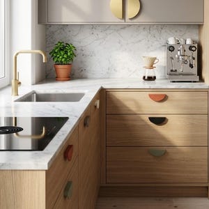 Può includere: Una cucina con armadi in legno chiaro e maniglie per cassetti semicircolari in vari colori, tra cui terracotta, nero e verde salvia. Lo spazio include un piano di lavoro in marmo bianco, un rubinetto dorato, un lavello e una macchina da caffè.