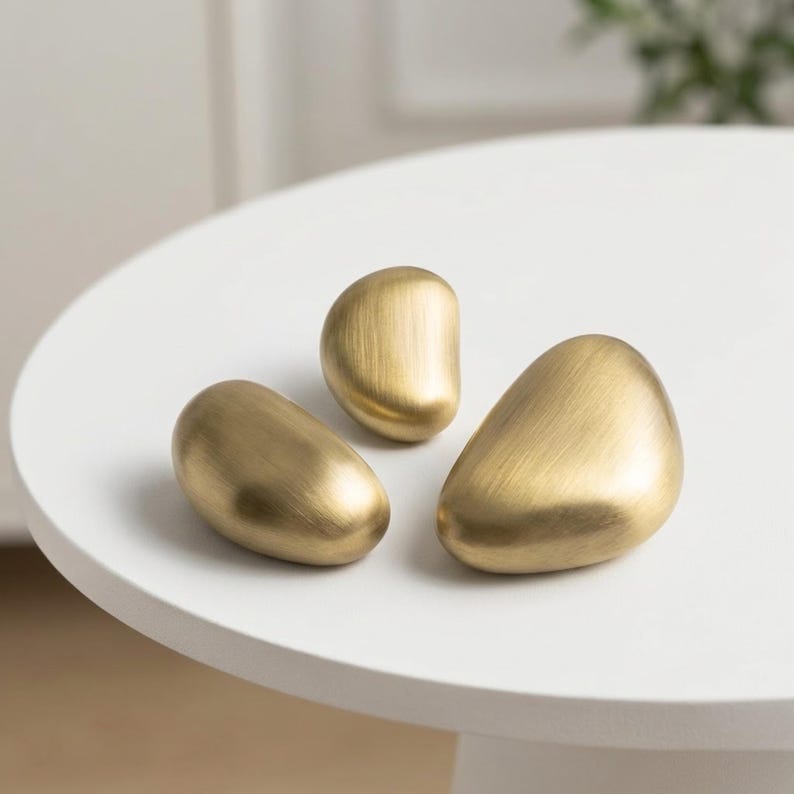 Op de afbeelding: Drie gladde, kiezelvormige objecten met een geborstelde gouden metalen afwerking. Elk stuk heeft een afgeronde, asymmetrische vorm met een zachte glans die het licht op het gebogen oppervlak reflecteert. De items zijn in een groep gerangschikt en tonen een consistente warme, gouden tint en een gepolijste, uniforme textuur over hun massieve, afgeronde lichamen.