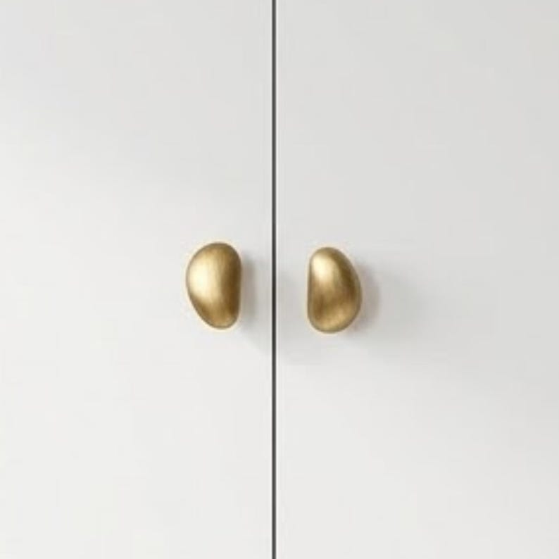 Op de afbeelding: Twee asymmetrische, kiezelvormige kastknoppen met een geborstelde gouden afwerking. Elke knop heeft een glad, afgerond oppervlak en is gemonteerd op een vlak, effen wit kastdeurpaneel, wat zorgt voor een strak en eenvoudig visueel contrast.