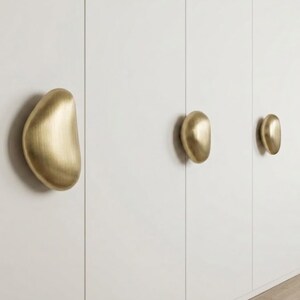 Op de afbeelding: Drie geborsteld messing kastknoppen met een organische, kiezelachtige vorm, gemonteerd op een wit oppervlak. Elke knop heeft een gladde, afgeronde, asymmetrische vorm met een metallic afwerking die het licht reflecteert voor een warme gloed.
