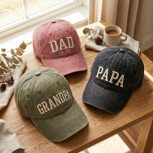 May include: Three baseball caps in dusty rose, olive green, and charcoal gray. Each cap has white embroidered text: "DAD EST. 1993", "GRANDPA", and "PAPA". The caps are displayed on a wooden surface near a window.