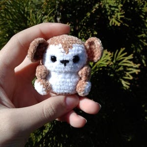 May include: A small, handmade crochet monkey with brown and white yarn. The monkey has large, round ears, black eyes, and small arms and legs. The plush toy is held in a hand against a blurred green background.