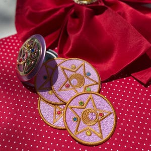 Op de afbeelding: Een ronde paarse geborduurde patch met een gouden ster- en maansikkelontwerp met kleine gekleurde accenten, vergezeld door een metallic goud en paars stervormig accessoire met ingelegde edelstenen, liggend op een rode stof met witte stippen.