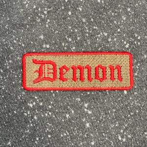 May include: A rectangular embroidered patch featuring the word Demon in red gothic-style lettering on a tan background, bordered by a red stitched edge. The patch is attached to a dark gray fabric with a white speckled or acid-wash pattern.