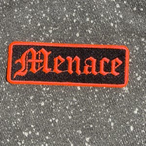 May include: A rectangular embroidered patch featuring the word Menace in a red gothic-style font. The patch has a black background and a thick red border, set against a dark gray fabric with a white speckled pattern throughout the material.