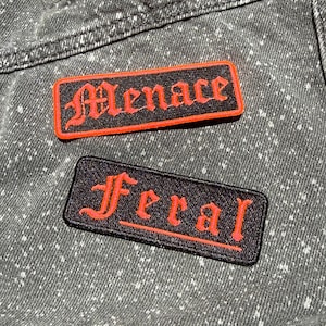 May include: Two rectangular embroidered patches on a speckled black denim fabric. The top patch features the word Menace in red gothic lettering with a red border. The bottom patch features the word Feral in red gothic lettering with a red border.
