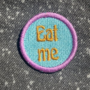 May include: A circular embroidered patch featuring a light blue background with a purple stitched border. The text "Eat me" is embroidered in the center in a golden yellow thread using a rounded, playful font style on the fabric surface.