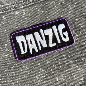 May include: A rectangular embroidered patch featuring the word DANZIG in white block lettering on a black background with a purple border, attached to a grey fabric garment with a white speckled, acid-wash style pattern throughout the material.