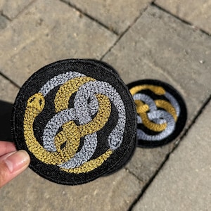 May include: Two circular embroidered patches featuring a black background with a design of two intertwined snakes, one in metallic gold thread and the other in silver thread, forming a continuous knot pattern with visible stitching texture.