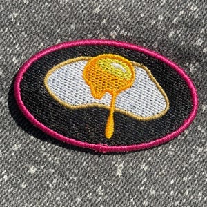 May include: An oval-shaped embroidered patch featuring a fried egg with a bright yellow yolk and white egg white on a black background. The patch is bordered by a vibrant pink stitched edge, creating a distinct contrast against the dark center.