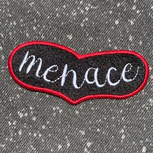 May include: An embroidered patch with a black heart-shaped base and a vibrant red border. The word menace is stitched across the center in white cursive script. The patch features a textured fabric surface with visible thread details throughout.