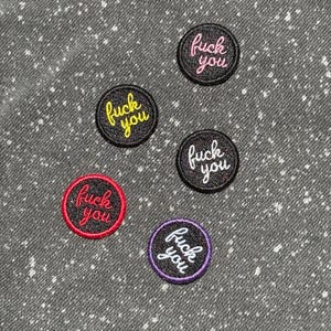 May include: Five circular embroidered patches featuring the text "fuck you" in cursive script. The patches have black backgrounds with borders and lettering in pink, yellow, white, red, and purple, arranged on a speckled gray fabric surface.