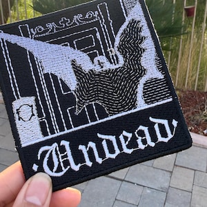 May include: A square black embroidered patch featuring a white stitched design of a bat with wings spread wide in front of a gate. The word Undead is embroidered in a gothic white font at the bottom of the patch with a stitched border edge.