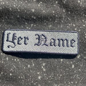 Könnte beinhalten: Ein rechteckiger gestickter Aufnäher mit hellgrauem Hintergrund, dunkelblauem Rand und dem Text "Yer Name", der in einer dunkelblauen gotischen Schrift gestickt ist. Der Aufnäher liegt auf einem dunkelgrauen, gesprenkelten Stoff.
