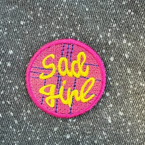 Può includere: Una toppa ricamata circolare con il testo "sad girl" in corsivo giallo. La toppa ha uno sfondo rosa con un motivo a griglia blu e un bordo rosa solido, il tutto applicato su un tessuto grigio scuro con macchioline bianche.