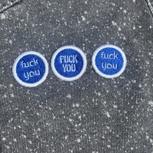 May include: A dark gray fabric with a white speckled pattern featuring three circular embroidered patches. Each patch has a blue background, a white border, and the text "fuck you" stitched in white thread across the center of the circle.