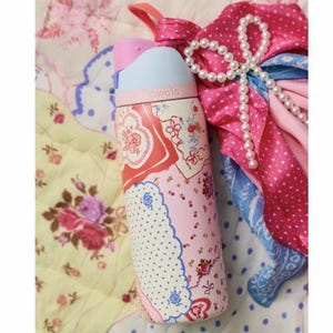 May include: A pink and white patterned water bottle with a light blue lid and a pink handle. The bottle has a floral and paisley design. The word "owala" is printed on the bottle. A pink scarf and a pearl necklace are next to the bottle.