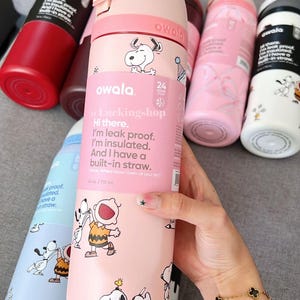 May include: Several Owala insulated water bottles in shades of pink, red, and blue. The pink bottle has Snoopy and Charlie Brown illustrations, with text: "Hi there. I'm leak proof. I'm insulated. And I have a built-in straw." The bottles are 710 ml.