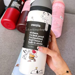 May include: Several Owala insulated water bottles in red, brown, pink, and light blue. The white bottle has Snoopy and Charlie Brown illustrations, with text: "Hi there. I'm leak proof. I'm insulated. And I have a built-in straw." The bottles are 945 ml.