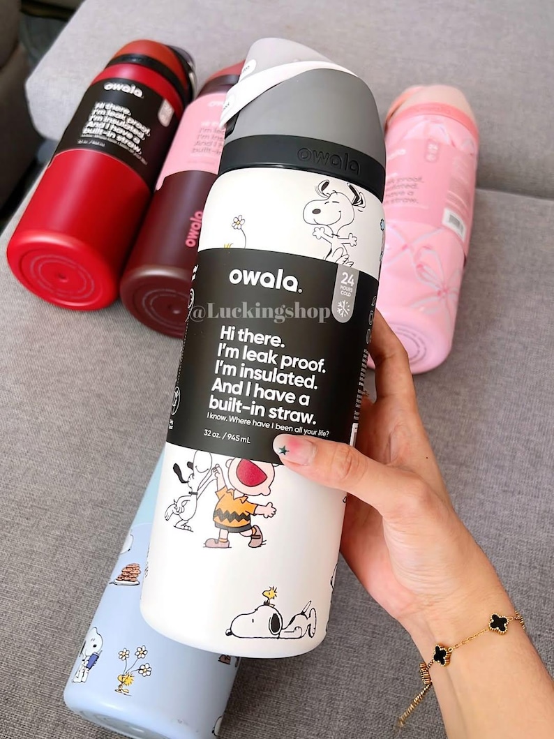 May include: Several Owala insulated water bottles in red, brown, pink, and light blue. The white bottle has Snoopy and Charlie Brown illustrations, with text: "Hi there. I'm leak proof. I'm insulated. And I have a built-in straw." The bottles are 945 ml.