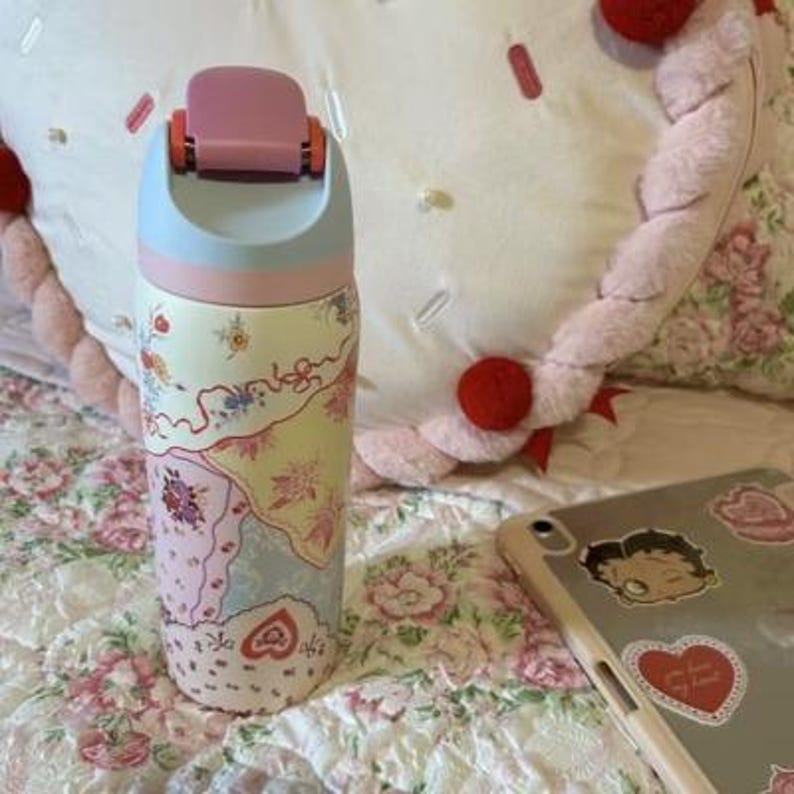 May include: A floral-patterned water bottle with a pink, blue, and cream color scheme. The bottle has a pink flip-top lid and a decorative design featuring flowers and hearts. The bottle is approximately 9 inches tall.
