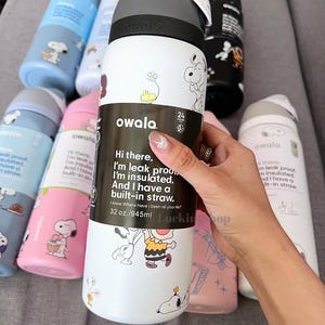 May include: White and black Owala insulated water bottle with a grey lid, featuring Snoopy and Charlie Brown graphics. The bottle has a built-in straw and holds 945ml. Other bottles in blue, pink, and black are visible.