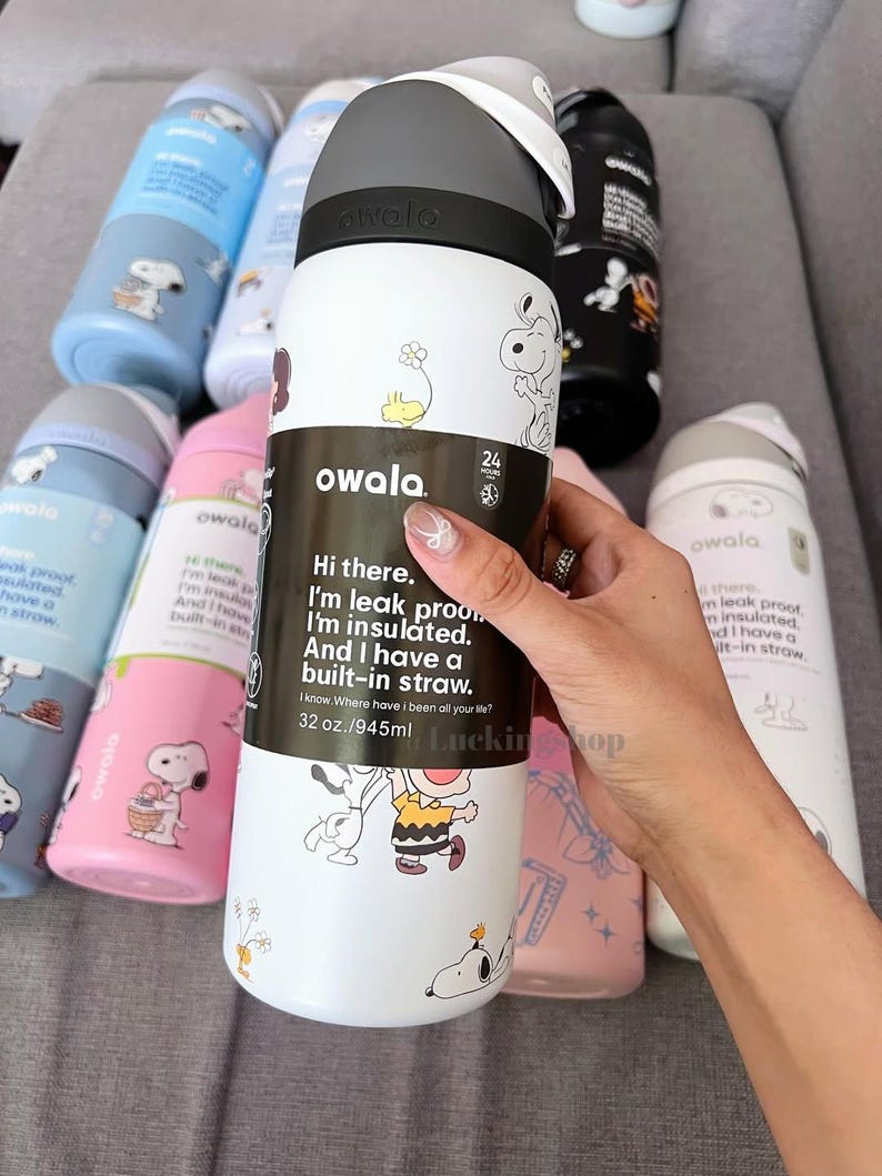 May include: White and black Owala insulated water bottle with a grey lid, featuring Snoopy and Charlie Brown graphics. The bottle has a built-in straw and holds 945ml. Other bottles in blue, pink, and black are visible.