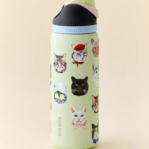 May include: A light green Owala water bottle featuring various cat faces with accessories. The bottle has a black and light green lid with a light blue band. The word "owala" is printed on the side.