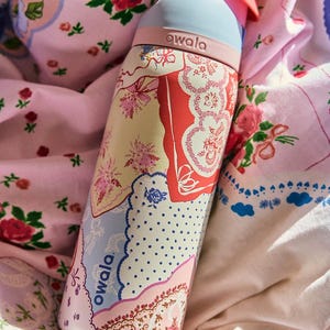May include: A floral patterned water bottle with a light blue top and pink accents. The bottle features a variety of floral designs in pink, red, and blue. The brand name "owala" is printed on the bottle.