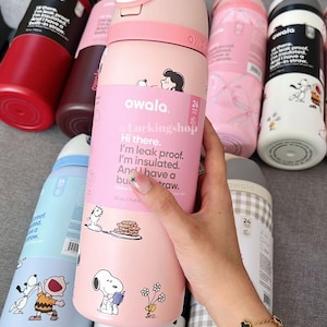 May include: Several Owala insulated water bottles in shades of pink, red, and blue. The bottles are decorated with Snoopy and Peanuts characters. The pink bottle in the center has the text "Hi there. I'm leak proof. I'm insulated. And I have a built-in straw."