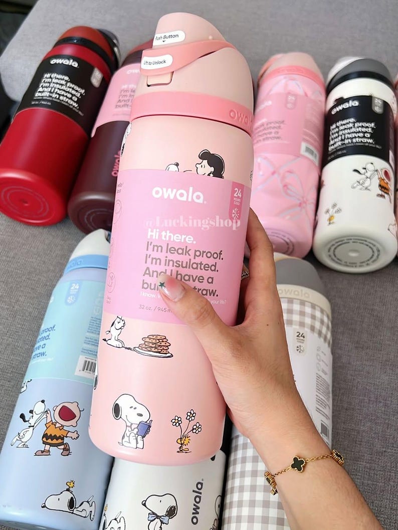May include: Several Owala insulated water bottles in shades of pink, red, and blue. The bottles are decorated with Snoopy and Peanuts characters. The pink bottle in the center has the text "Hi there. I'm leak proof. I'm insulated. And I have a built-in straw."
