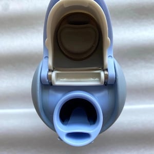 May include: A pale blue water bottle with a white flip-top lid. The bottle has a rounded shape with a wide opening at the bottom and a smaller spout. The lid is open, revealing the interior of the bottle.