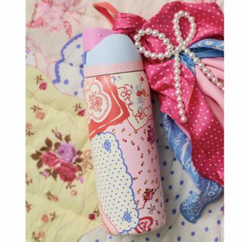 May include: A pink and white patterned water bottle with a light blue lid and a pink handle. The bottle has a floral and paisley design. The word "owala" is printed on the bottle. A pink scarf and a pearl necklace are next to the bottle.