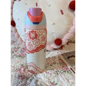 May include: A patterned water bottle with a light blue, pink, and orange lid. The body of the bottle features a floral design in red and white, with a dotted pattern at the bottom. The bottle is on a floral patterned bedspread.