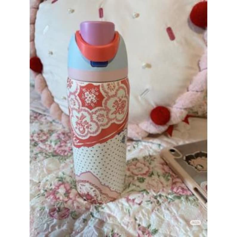 May include: A patterned water bottle with a light blue, pink, and orange lid. The body of the bottle features a floral design in red and white, with a dotted pattern at the bottom. The bottle is on a floral patterned bedspread.