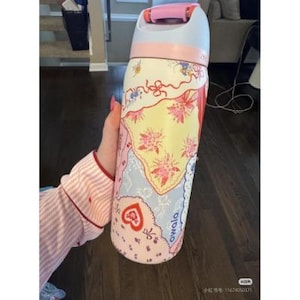 May include: A floral patterned water bottle with a pink and light blue lid. The bottle features a mix of pastel colors, including pink, yellow, and blue, with floral and heart designs. The word "owala" is printed on the side.