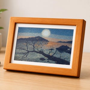 May include: A digital photo frame with a light brown wooden border displaying a landscape image of a lake at sunset with a tree in the foreground. The screen shows the time 12:30 and the date 27 Jul in white text centered over the water and mountains.