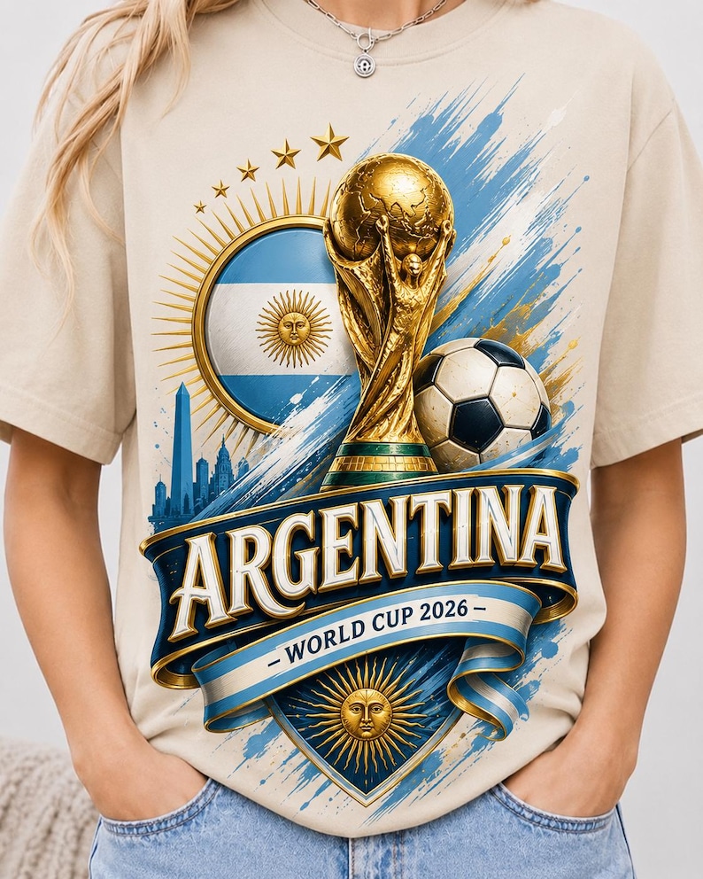 May include: A beige t-shirt featuring a graphic print of the gold World Cup trophy, a football, the Argentina flag, and the text "ARGENTINA - WORLD CUP 2026". The design includes blue and gold accents, a sun emblem, and a city skyline silhouette.