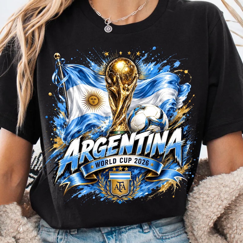 May include: A black t-shirt featuring a graphic print of the Argentina flag, a gold World Cup trophy, and a football. The text reads "ARGENTINA" and "WORLD CUP 2026" above the AFA crest, surrounded by blue and gold paint splatter effects.