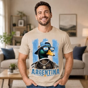 May include: A beige short-sleeve t-shirt featuring a graphic of a mallard duck wearing a blue baseball cap and sunglasses with the Argentina flag reflected in the lenses. The duck wears a silver chain, and the word ARGENTINA is printed below.