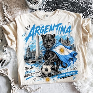 May include: A cream-coloured t-shirt featuring a graphic of a black panther draped in an Argentina flag, standing over a football. The design includes the text "ARGENTINA," "PASI&Oacute;N ORGULLO ALBICIELESTE," and "&iexcl;VAMOS ARGENTINA!" with city landmarks.