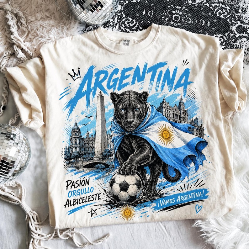 May include: A cream-coloured t-shirt featuring a graphic of a black panther draped in an Argentina flag, standing over a football. The design includes the text "ARGENTINA," "PASI&Oacute;N ORGULLO ALBICIELESTE," and "&iexcl;VAMOS ARGENTINA!" with city landmarks.